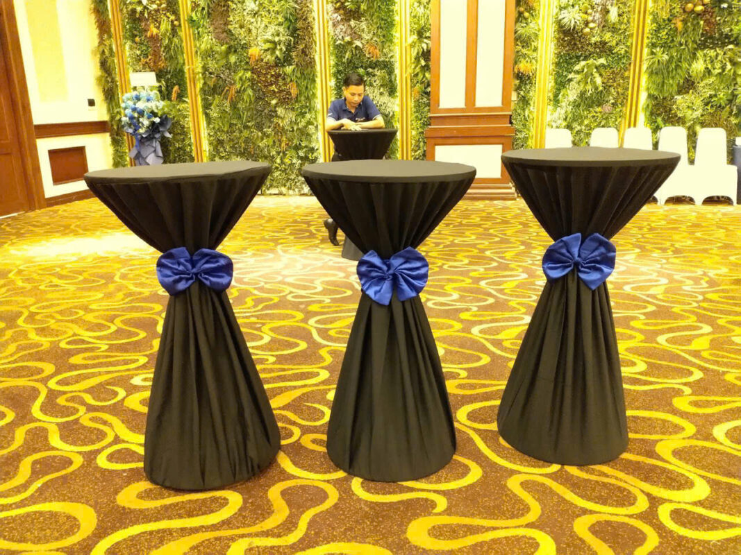 Bàn cocktail phủ khăn đen thắt nơ xanh trong sảnh trải thảm